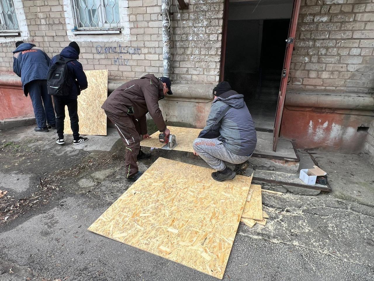 Робітники на вулиці біля під'їзду житлового будинку розпилюють листи OSB-плит за допомогою дискової пилки. Вони готують матеріали для закриття пошкоджених вікон або дверних отворів після обстрілу.