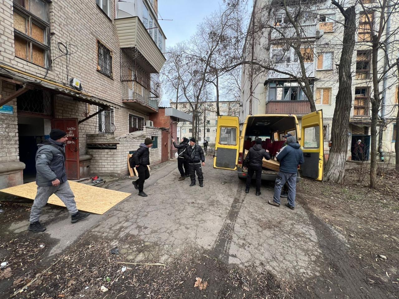 На подвір'ї багатоповерхового будинку стоїть жовтий мікроавтобус із відчиненими задніми дверцятами, з якого робітники вивантажують будівельні матеріали. На задньому плані видно житловий будинок, де частина вікон уже забита плитами OSB. Група людей координує роботу з відновлення будівлі.