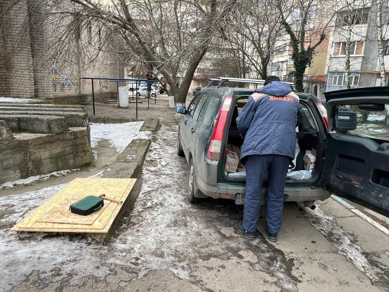 На засніженому подвір'ї житлового масиву робітник у спецодязі розвантажує інструменти з багажника позашляховика. Поруч на землі лежать плити OSB та будівельне приладдя, підготовлені для проведення ремонтних робіт. На задньому плані видно багатоповерхові будинки з вікнами, закритими захисними щитами.