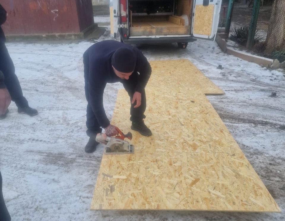 На засніженій ділянці чоловік у темному одязі розпилює великий лист OSB-плити за допомогою ручної циркулярної пилки. Позаду нього стоїть білий мікроавтобус із відчиненими задніми дверцятами, в якому складено будівельні матеріали.