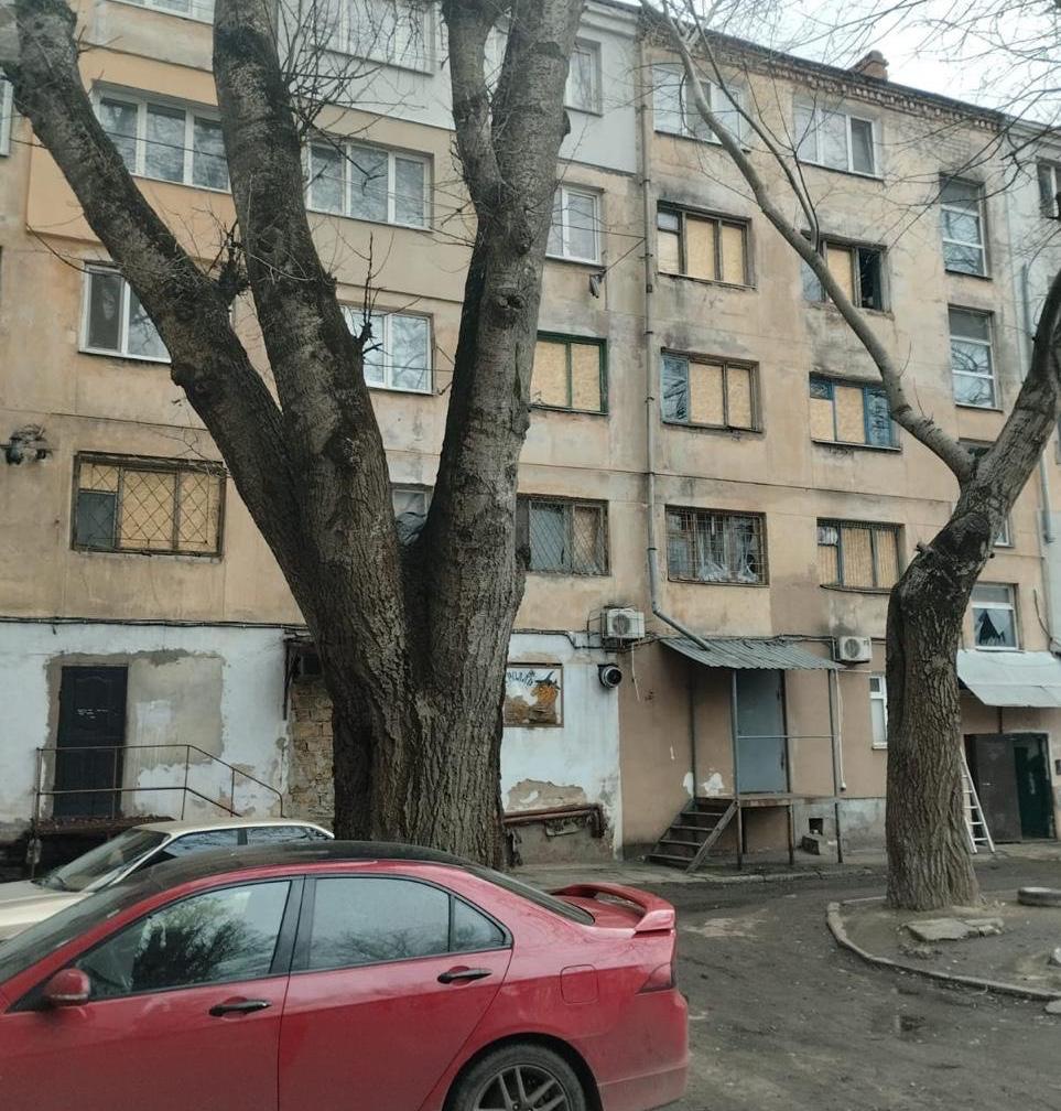 Фасад багатоповерхового житлового будинку, пошкодженого внаслідок обстрілу. Багато вікон на різних поверхах розбиті або вже закриті плитами OSB. На передньому плані видно припарковані автомобілі та голі дерева.