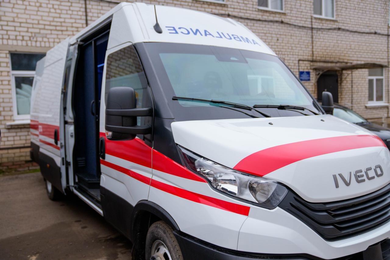 Білий мікроавтобус Iveco, переобладнаний під швидку допомогу, стоїть під кутом біля цегляної будівлі. На капоті та над лобовим склом помітний напис «AMBULANCE» у дзеркальному відображенні та червона декоративна смуга. Бічні двері автомобіля відчинені.