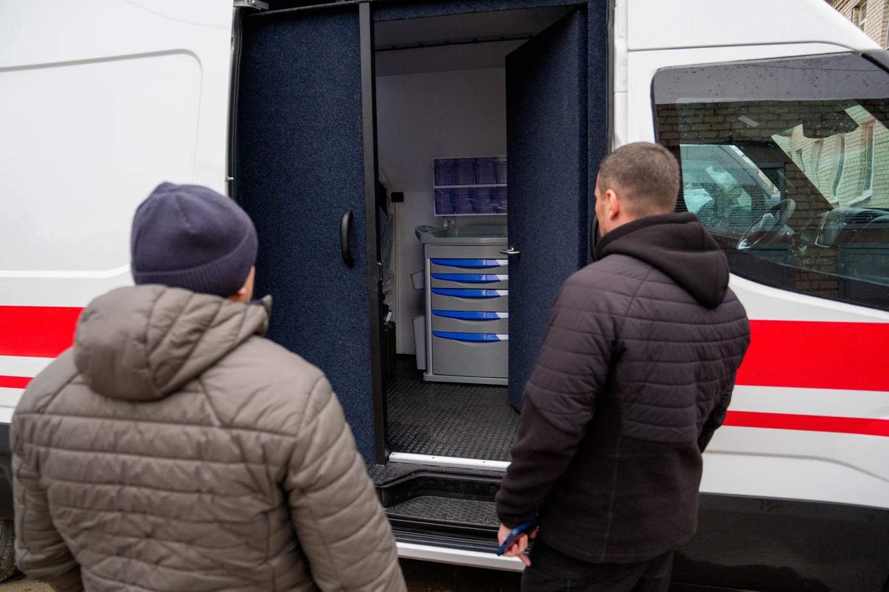 Двоє чоловіків зі спини розглядають внутрішнє оснащення медичного автомобіля через відкриті бічні двері. Всередині кабіни видно сірий медичний візок із синіми шухлядами та органайзер для препаратів. Автомобіль білого кольору з червоною горизонтальною смугою на кузові.
