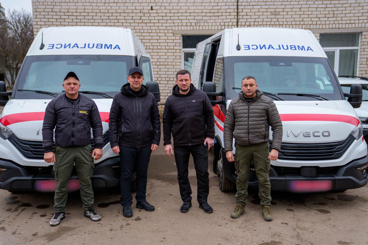 Чотири чоловіки стоять перед двома білими каретами швидкої допомоги марки Iveco. Автомобілі мають характерне червоне маркування та напис «AMBULANCE», виконаний у дзеркальному відображенні на капоті. Чоловіки одягнені у повсякденний та тактичний одяг темних кольорів, на фоні видно цегляну будівлю.