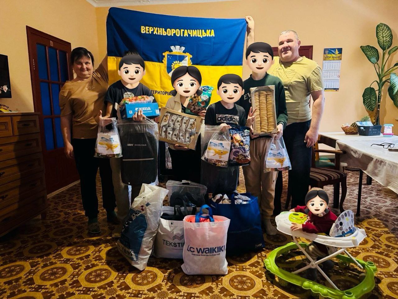 Група дорослих і дітей у кімнаті тримають прапор Верхньорогачицької громади та численні пакунки з гуманітарною допомогою, солодощами та речами. Поруч на підлозі стоять великі сумки з продуктами та засобами, а на передньому плані дитина сидить у дитячих ходунках.