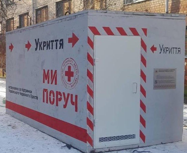 Наземне бетонне укриття сірого кольору, встановлене на вулиці. На стіні споруди нанесено написи «Укриття» зі стрілками, гасло «МИ ПОРУЧ» та емблема Червоного Хреста України. Двері укриття по периметру позначені червоно-білими світловідбивними смугами.