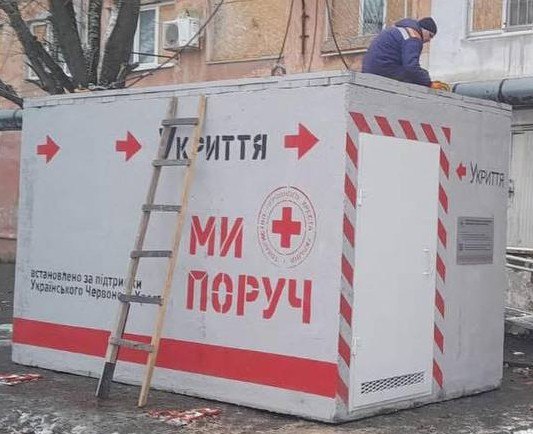 Наземне бетонне укриття білого кольору з написом «Укриття» та червоними стрілками-покажчиками. На стіні також розміщено логотип Червоного Хреста та напис «Ми поруч». Біля споруди стоїть дерев’яна драбина, а зверху на даху працює людина у спецодязі.