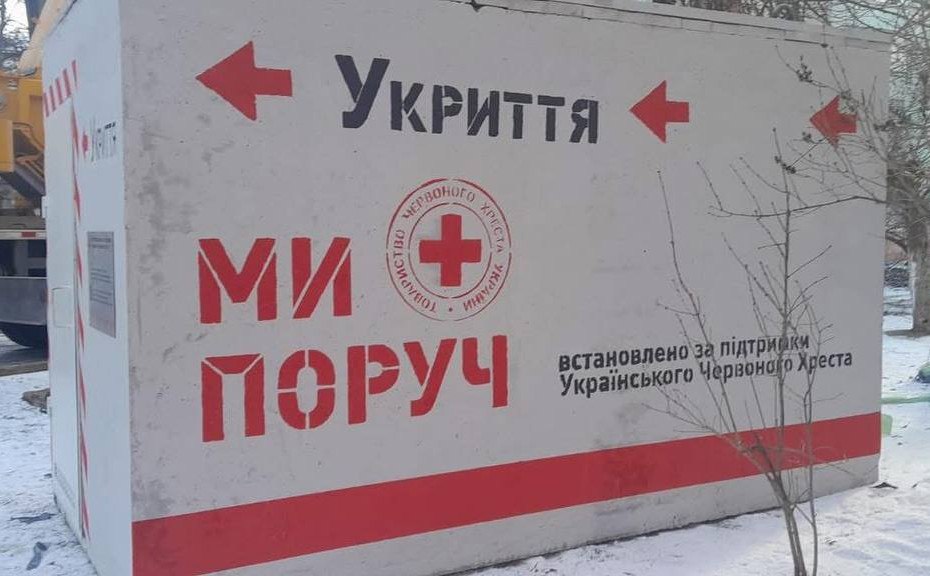 Біле бетонне мобільне укриття, встановлене на засніженій ділянці. На стіні великими літерами написано «Укриття» та «Ми поруч» поруч із емблемою Червоного Хреста. Також присутні червоні стрілки-покажчики та текст про те, що споруду встановлено за підтримки Українського Червоного Хреста.