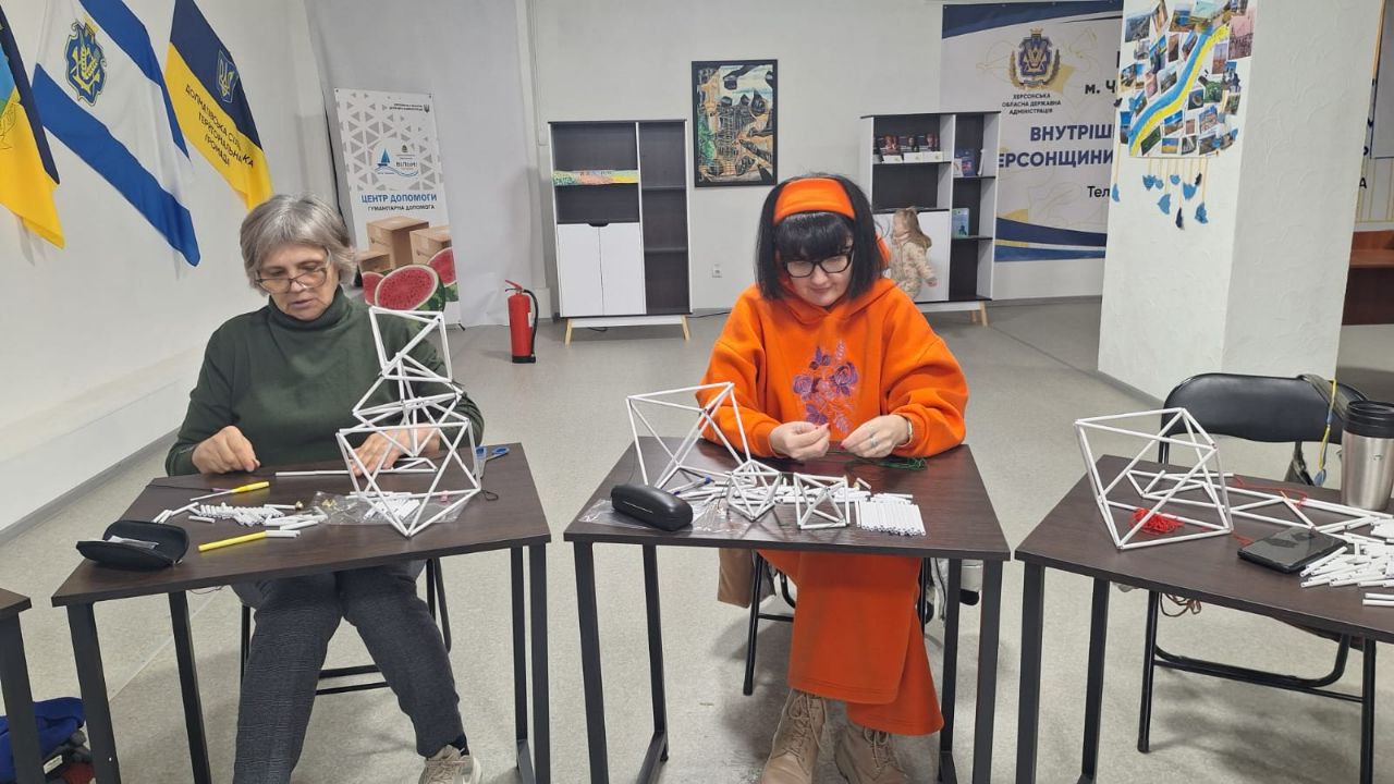 Дві жінки сидять за столами у світлому приміщенні та виготовляють об'ємні геометричні фігури з білих соломинок. На столах розкладені матеріали для творчості, а на задньому плані видно прапори, інформаційні стенди та меблі. Жінки зосереджені на процесі рукоділля.