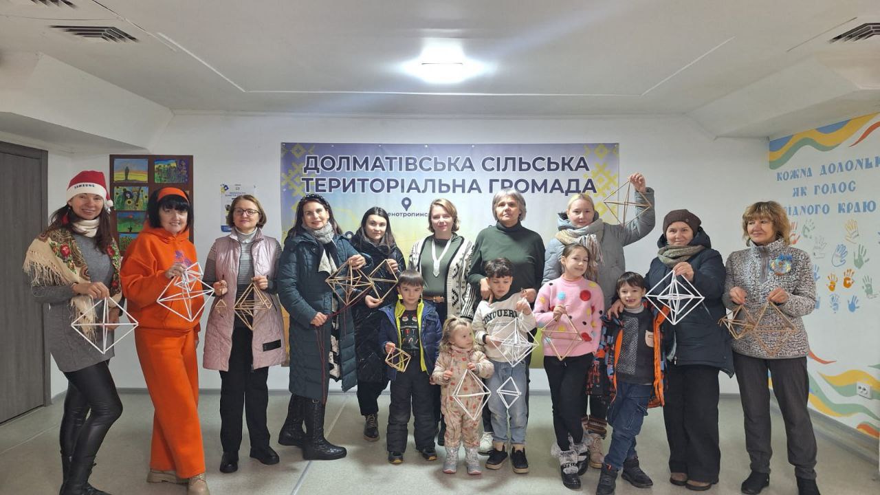 Група дорослих та дітей позикують у приміщенні на фоні банера з написом «Долматівська сільська територіальна громада». Учасники тримають у руках традиційні солом’яні вироби геометричної форми (різдвяні павуки). Приміщення світле, прикрашене малюнками та плакатами на стінах.
