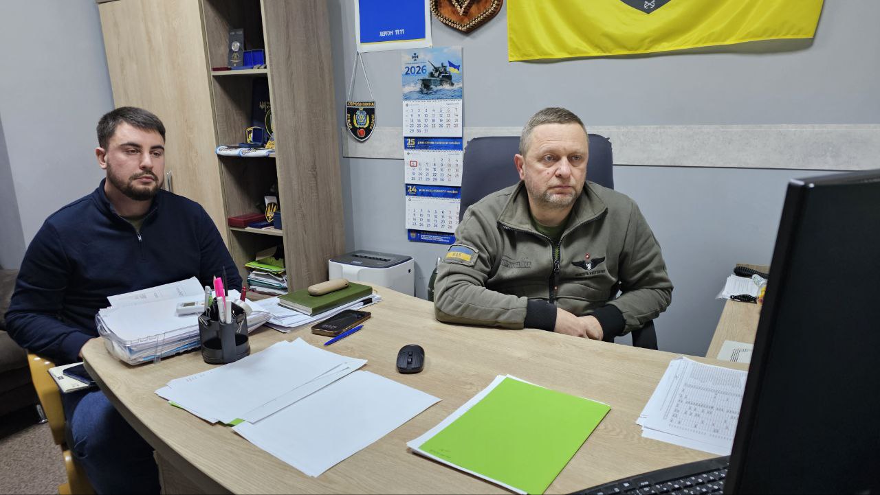 Двоє чоловіків сидять за робочим столом в офісі. Один із них у тактичному одязі, інший — у темно-синьому джемпері. На стіні за ними висить календар на 2026 рік, українська символіка та прапор.