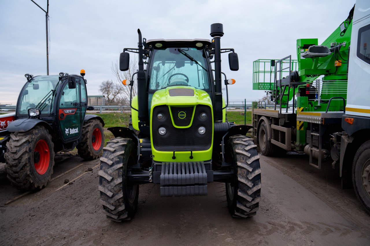 Яскраво-салатовий трактор стоїть на відкритому майданчику. Поряд з ним розташована інша спецтехніка: ліворуч — компактний навантажувач Bobcat, праворуч — частина білої вантажівки з краном-маніпулятором. Техніка розміщена на ґрунтовій поверхні під похмурим небом.