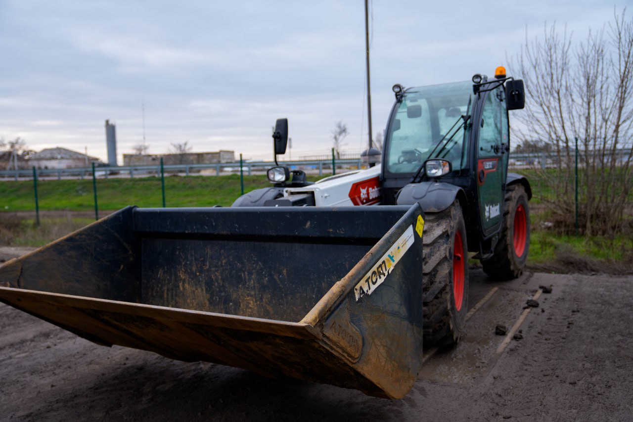 Компактний телескопічний навантажувач Bobcat з великим переднім ковшем стоїть на відкритому майданчику. Машина має чорно-білу кабіну та характерні червоні диски коліс. На ковші видно маркування, техніка розміщена на ґрунтовому покритті під хмарним небом.