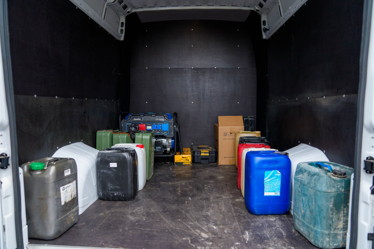 Внутрішній простір вантажного мікроавтобуса, заповнений обладнанням та каністрами. У глибині стоїть бензиновий генератор, поруч — картонна коробка та інструментальний ящик. На підлозі вздовж бортів розміщено багато пластикових та металевих каністр різних кольорів.
