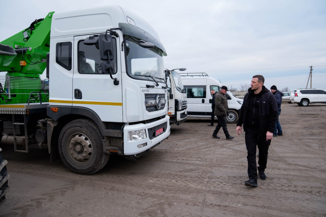 На передньому плані чоловік у темному спортивному одязі та чорній куртці йде по майданчику повз велику білу вантажівку з зеленим підйомним механізмом. Позаду нього на відкритому просторі розташовані інші транспортні засоби та кілька людей, що стоять групами. Техніка та персонал знаходяться на ґрунтовій поверхні під хмарним небом.