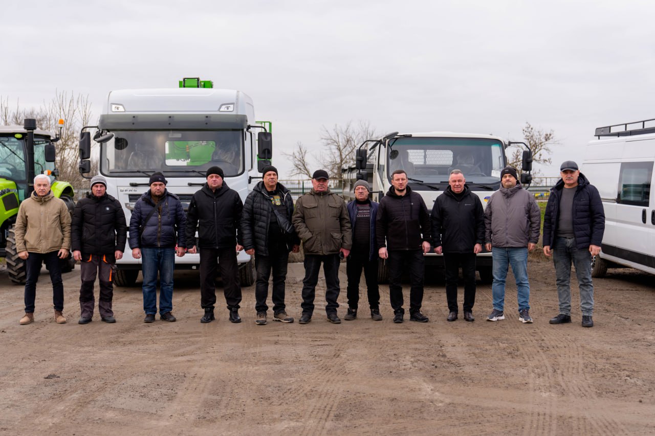 Група з одинадцяти чоловіків у теплому повсякденному та робочому одязі стоїть в один ряд на відкритому майданчику. Позаду них вишикувана спецтехніка: салатовий трактор, дві білі вантажівки (одна з зеленим підйомником) та білий мікроавтобус. Усі люди та техніка розміщені на ґрунтовій поверхні під хмарним небом.