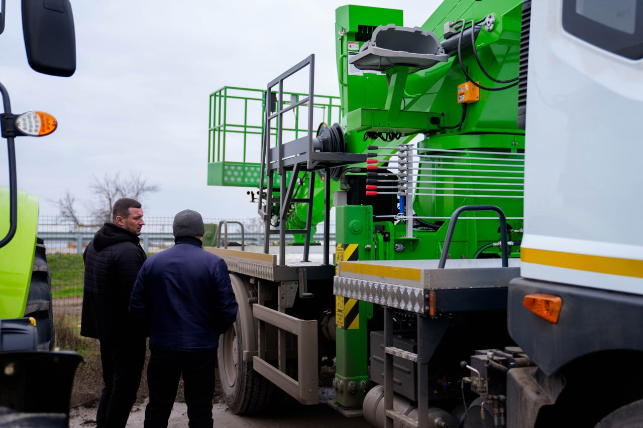 Двоє чоловіків стоять біля великої вантажівки, обладнаної зеленим краном-маніпулятором та підйомною платформою. На задньому плані видно металеві важелі керування та гідравлічні системи установки. Техніка розташована на відкритому майданчику в похмуру погоду.