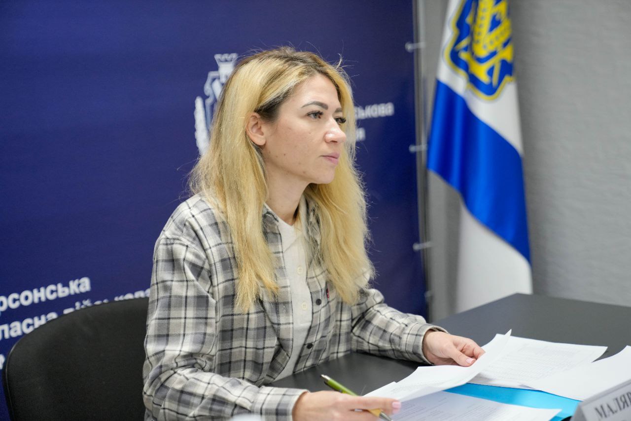 Жінка в картатій сорочці сидить за столом з паперами в руках, уважно слухаючи співрозмовника. Позаду неї розташований темно-синій брендований фон та державний прапор.