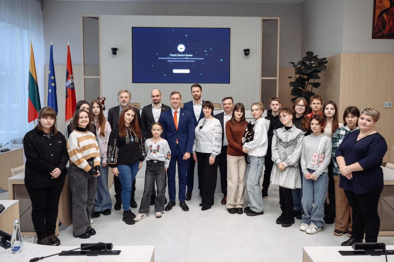 Групова фотографія делегації дорослих та молоді у світлій сучасній залі засідань. Позаду групи розміщено великий екран та прапори Литви, Європейського Союзу та ще один офіційний стяг. Учасники стоять у кілька рядів, деякі одягнені у вишиванки.