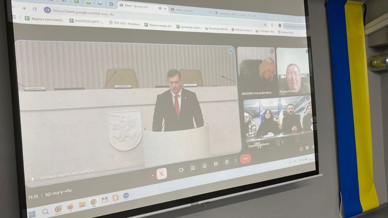 Проєкційний екран у приміщенні, на якому відображено вікно відеоконференції. На основному кадрі видно доповідача за трибуною в офіційній залі, а збоку — кілька вікон з іншими учасниками онлайн-зустрічі. Справа від екрана на стіні закріплено державний прапор України.