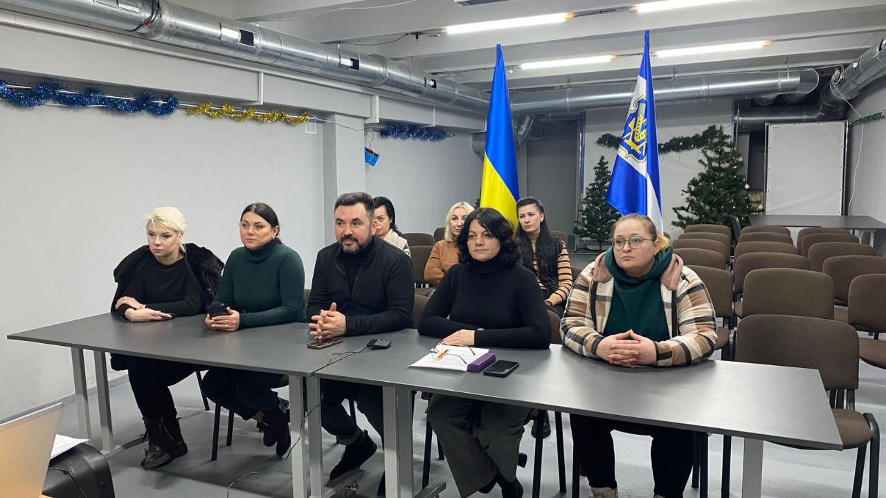 Група людей сидить за довгим столом під час офіційної зустрічі або наради у приміщенні, що нагадує облаштоване укриття. На задньому плані встановлені державний прапор України та прапор із гербом області, а стіни оздоблені скромним новорічним декором.