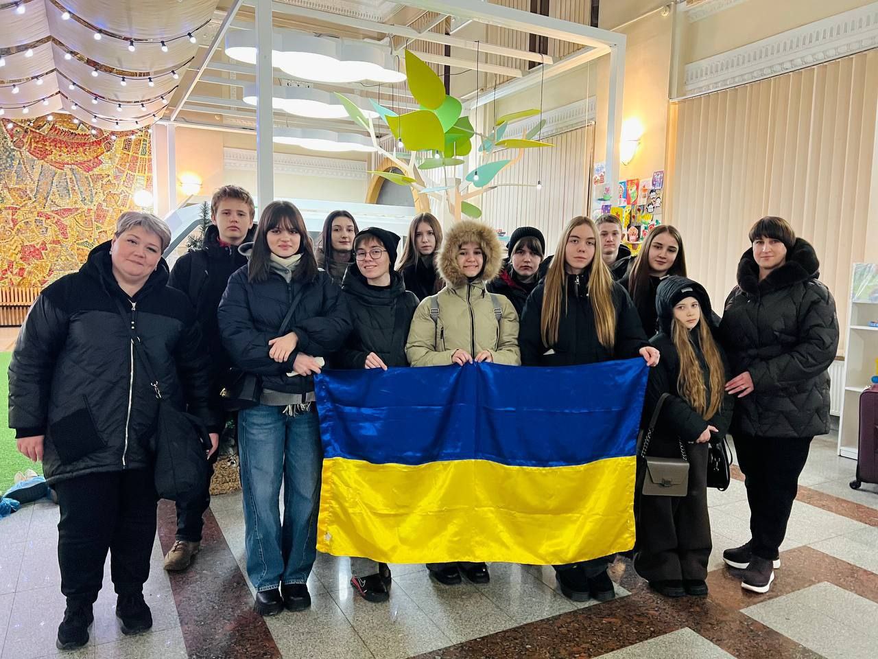 Групова фотографія дітей та дорослих у світлому залі. У центрі групи підлітки тримають розгорнутий державний прапор України. Приміщення прикрашене святковою ілюмінацією та декоративним деревом, а на задньому плані видно мозаїчне панно на стіні.