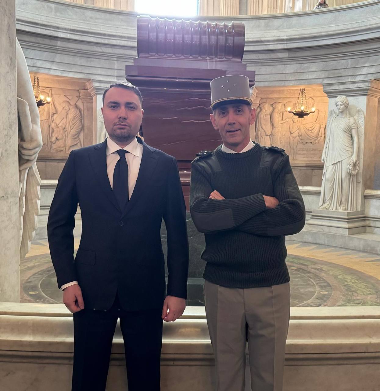 Спільна фотографія двох чоловіків у величному круглому залі з класичною архітектурою, колонами та античними скульптурами. Один чоловік одягнений у класичний темний костюм із краваткою, інший — у військову форму іноземного зразка з характерним головним убором. На задньому плані по центру розташований великий монумент або саркофаг із темного каменю.