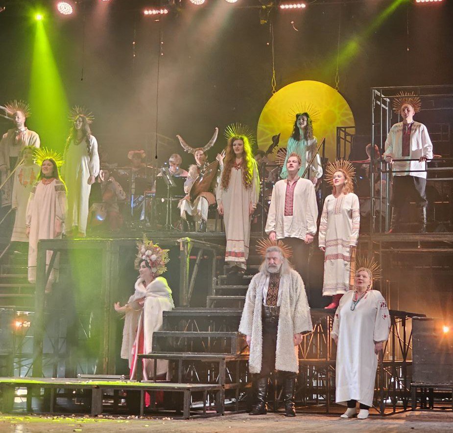 Актори у світлому етнічному вбранні на театральній сцені під час вистави. У центрі — велике жовте коло, що символізує сонце. Декорації мають вигляд багаторівневих металевих конструкцій, сцена яскраво освітлена прожекторами з жовтими та зеленими променями.