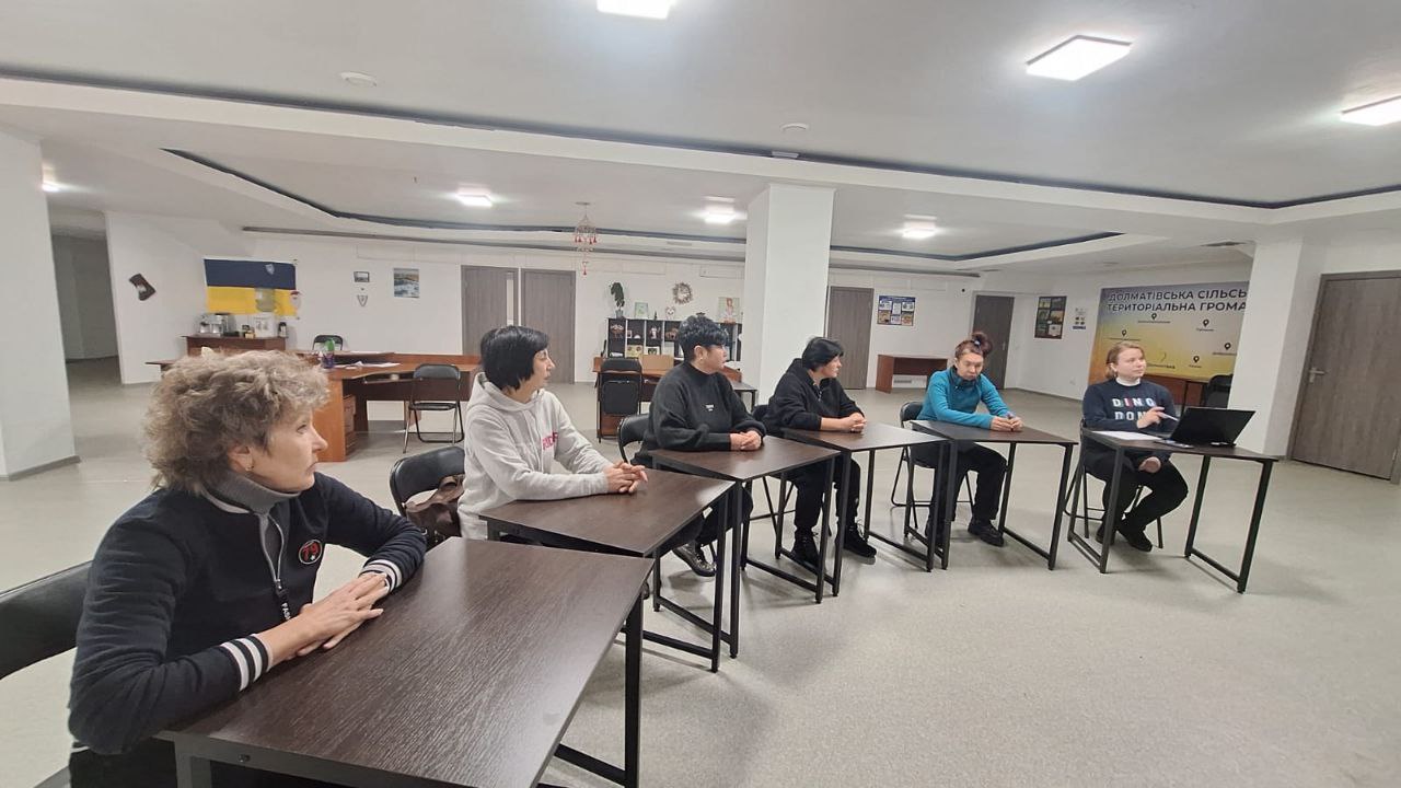 Група жінок сидить за окремими столами у просторому світлому офісі під час робочої зустрічі або тренінгу. Одна з учасниць працює за ноутбуком, інші уважно слухають. На фоні видно каву-зону, державну символіку та інформаційні стенди територіальних громад.