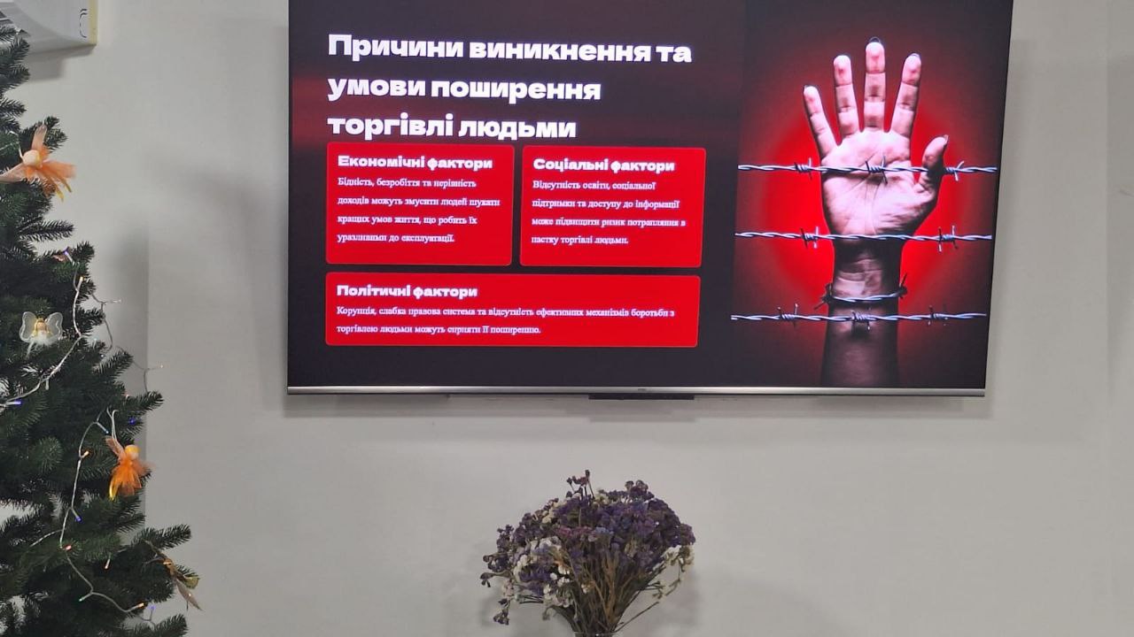 Екран монітора у світлому приміщенні, на якому відображено слайд презентації на тему причин виникнення торгівлі людьми. Текст на слайді розбитий на три блоки: економічні, соціальні та політичні фактори. Праворуч на екрані зображено руку за колючим дротом на червоному фоні. Поруч із монітором видно частину прикрашеної новорічної ялинки та вазу з квітами.