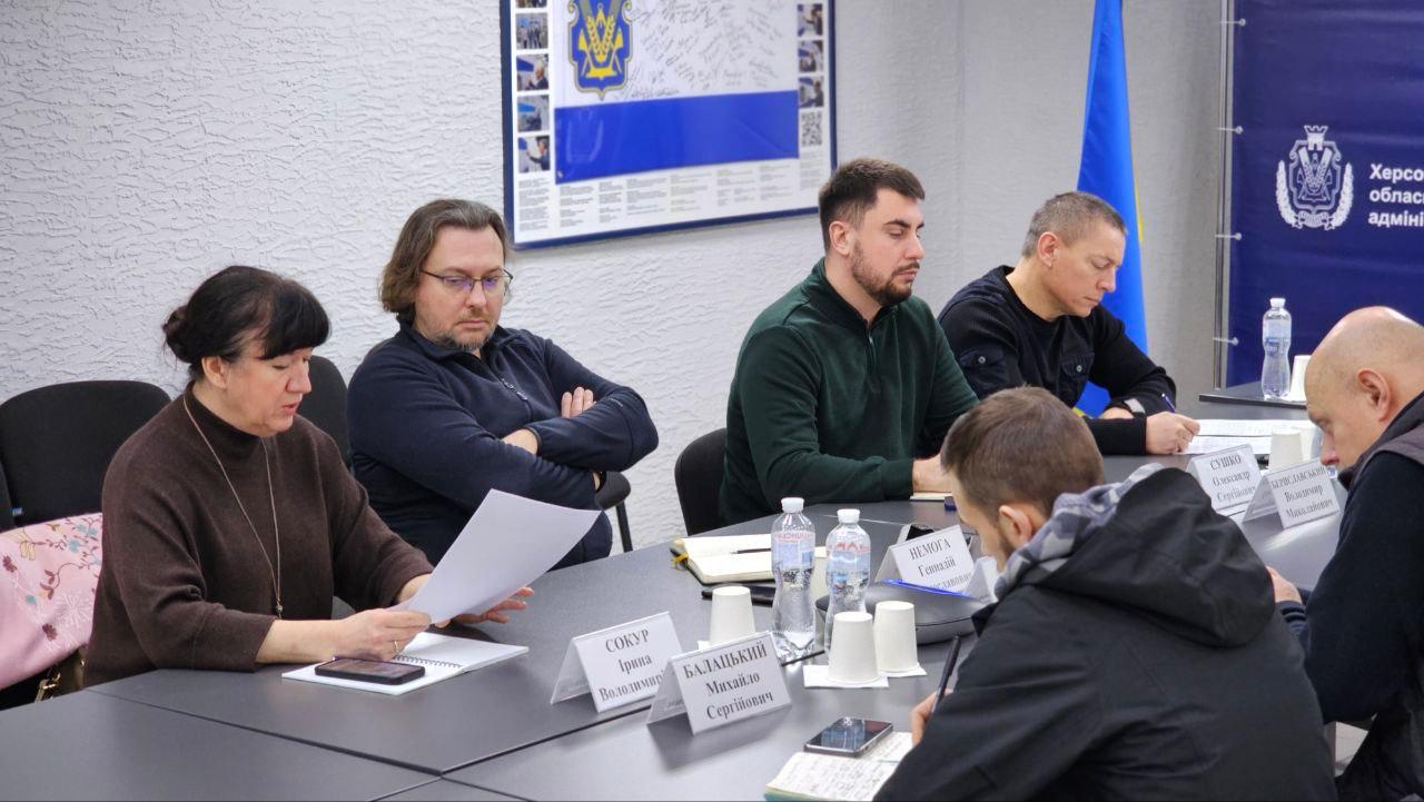 Учасники робочої наради сидять за столом у кабінеті. На передньому плані жінка переглядає документи, поруч сидять чоловіки, які уважно слухають або роблять нотатки. Перед кожним учасником стоять таблички з іменами та пляшки з водою. На фоні видно частину інформаційного стенда з державним гербом та синій банер.