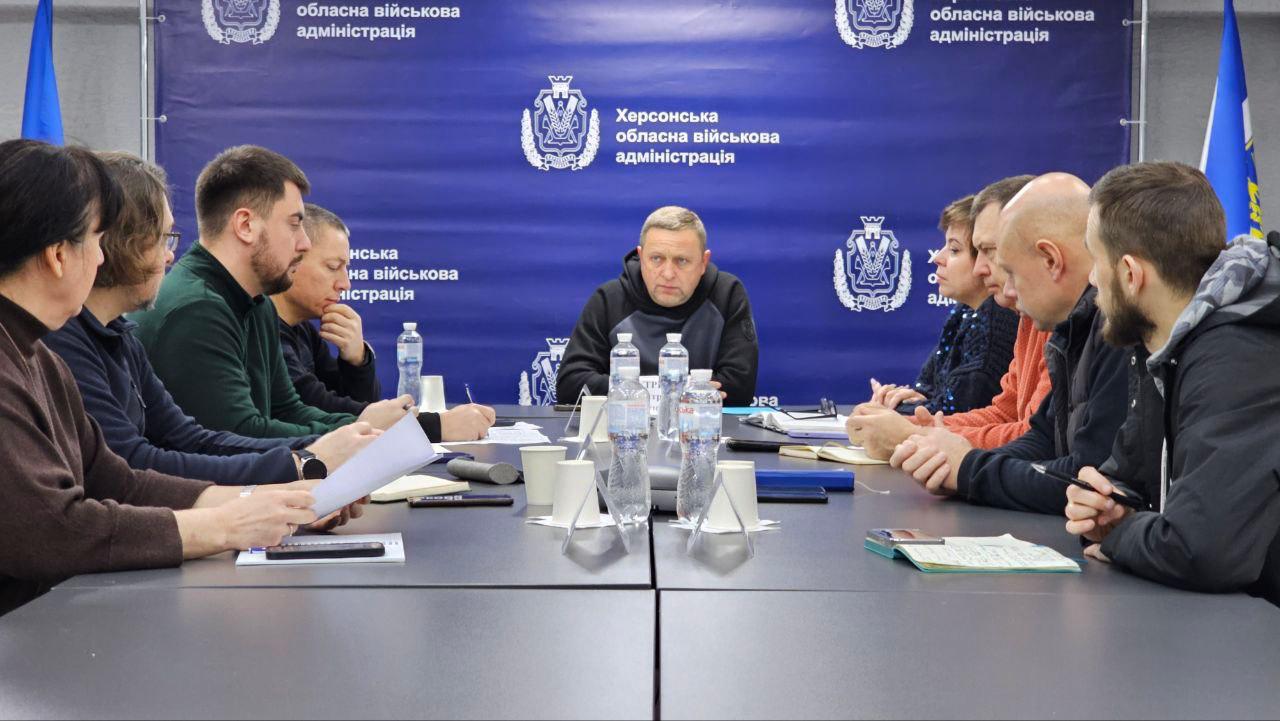 Робоча нарада за великим столом у приміщенні з офіційною символікою. Група людей сидить один навпроти одного, обговорюючи робочі питання. На фоні розміщено синій банер із логотипами Херсонської обласної військової адміністрації, на столі стоять пляшки з водою, блокноти та мобільні телефони.