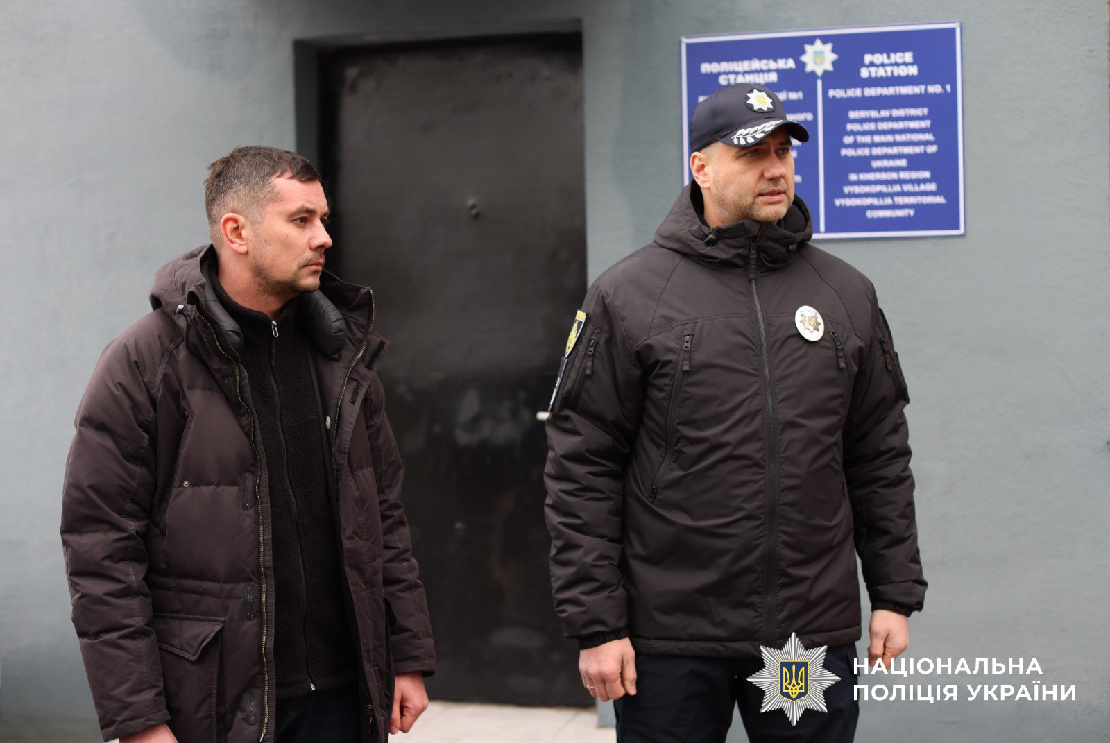Двоє чоловіків — один у цивільному верхньому одязі, інший у темній куртці з жетоном поліції та кепці з емблемою — стоять на фоні входу до будівлі з інформаційною табличкою «Поліцейська станція». У правому нижньому куті розміщено логотип Національної поліції України.