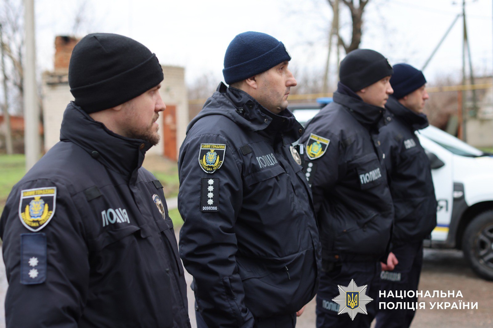 Четверо співробітників поліції у темному зимовому однострої та шапках стоять у шерензі (вид збоку). На рукавах курток видно шеврони Національної поліції. На задньому плані стоїть службовий автомобіль, а в нижньому куті розміщено логотип відомства.