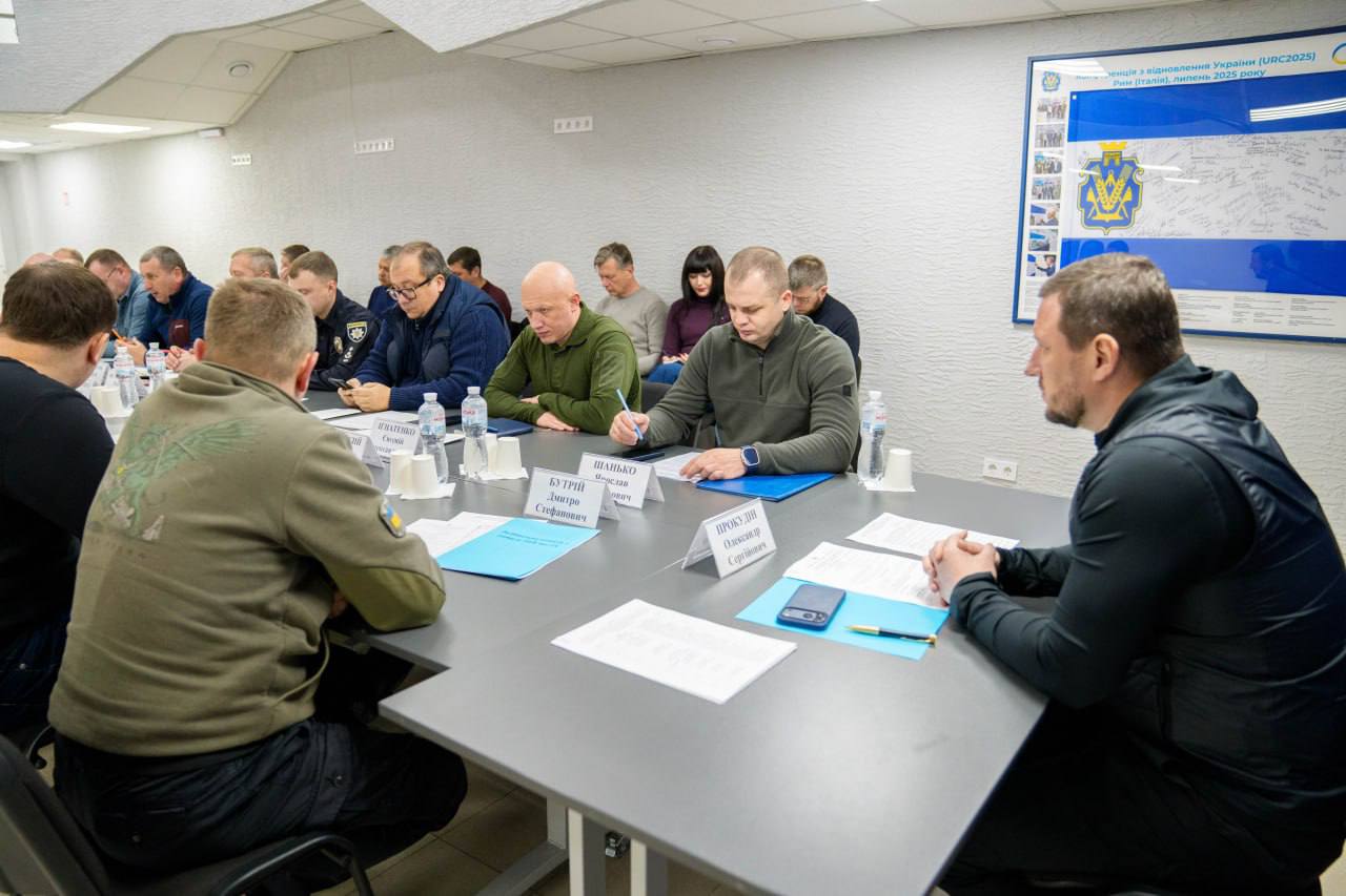 Нарада за довгим столом у конференц-залі. Учасники в офіційному та мілітарі-одязі вивчають документи та роблять нотатки. На столі розміщені таблички з прізвищами, пляшки з водою та папери. На стіні праворуч висить інформаційний стенд із символікою та текстом.