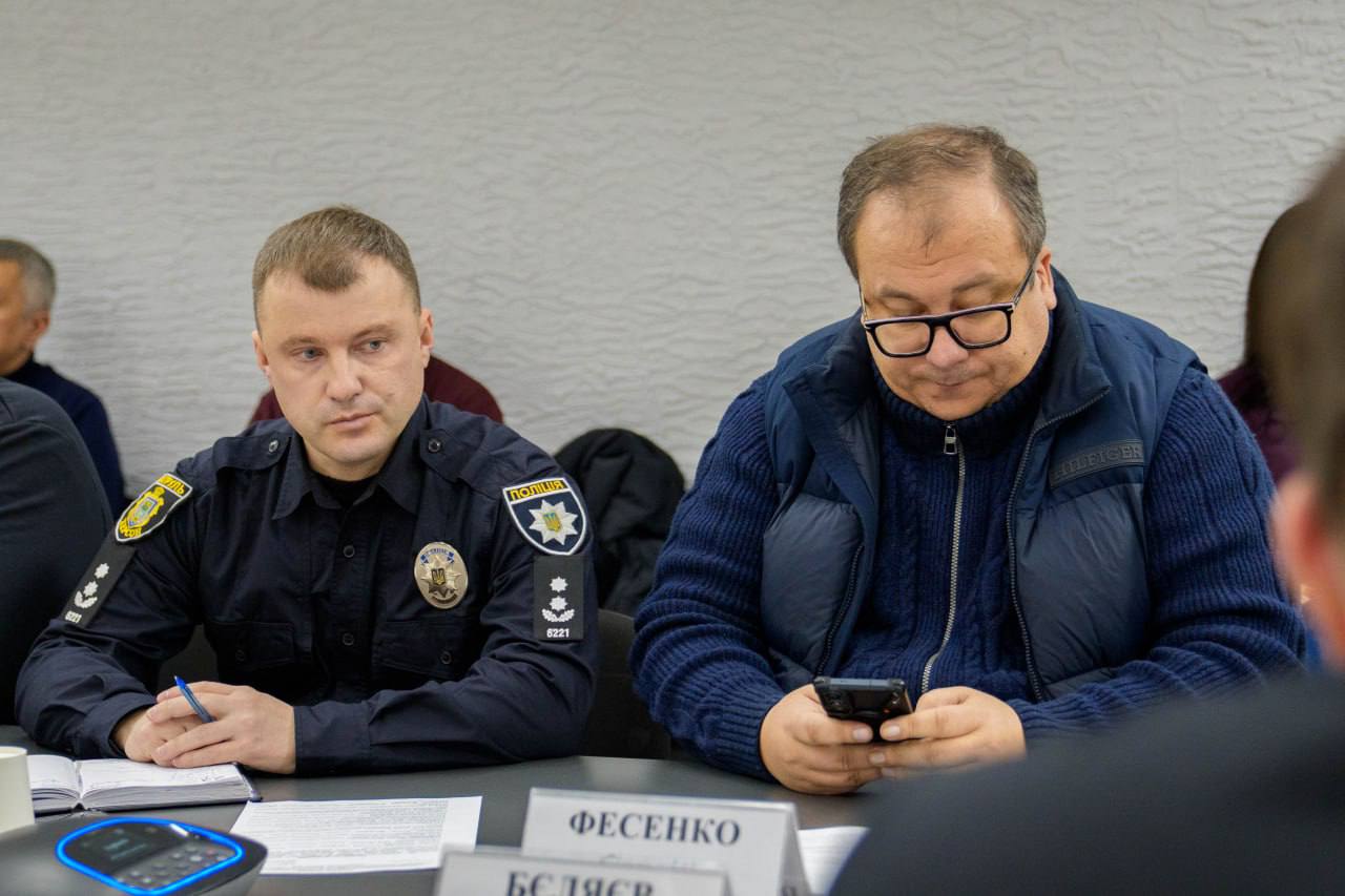 Двоє чоловіків за робочим столом під час офіційного заходу. Ліворуч — співробітник поліції в темному однострої з шевронами, який тримає ручку. Праворуч — чоловік у синьому в'язаному жилеті, який дивиться в телефон.