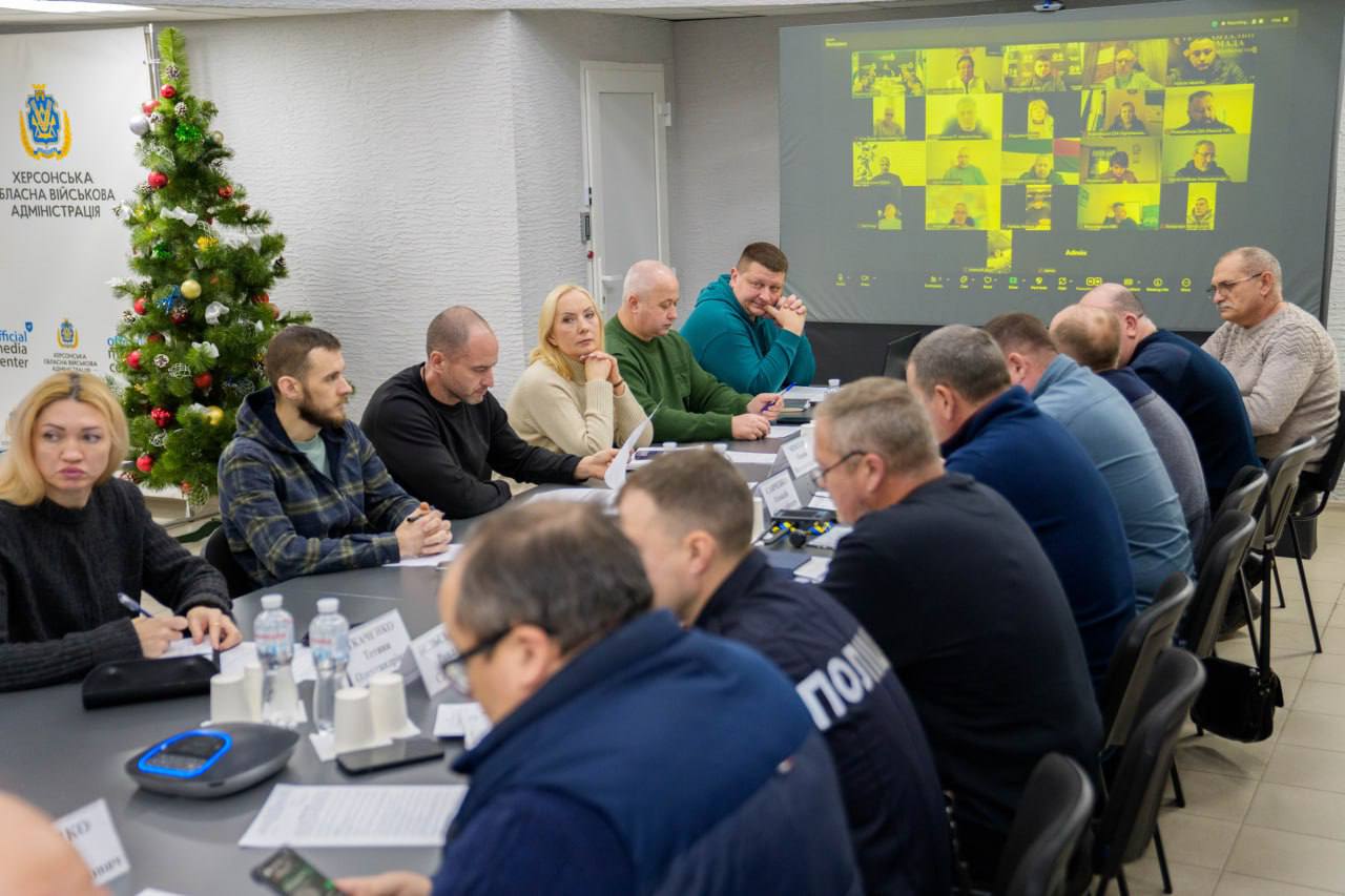 Учасники офіційної наради сидять за великим столом у конференц-залі. На передньому плані ліворуч стоїть прикрашена новорічна ялинка. У центрі кімнати встановлено великий екран, на якому транслюється відеозв'язок з іншими учасниками у форматі онлайн-конференції.