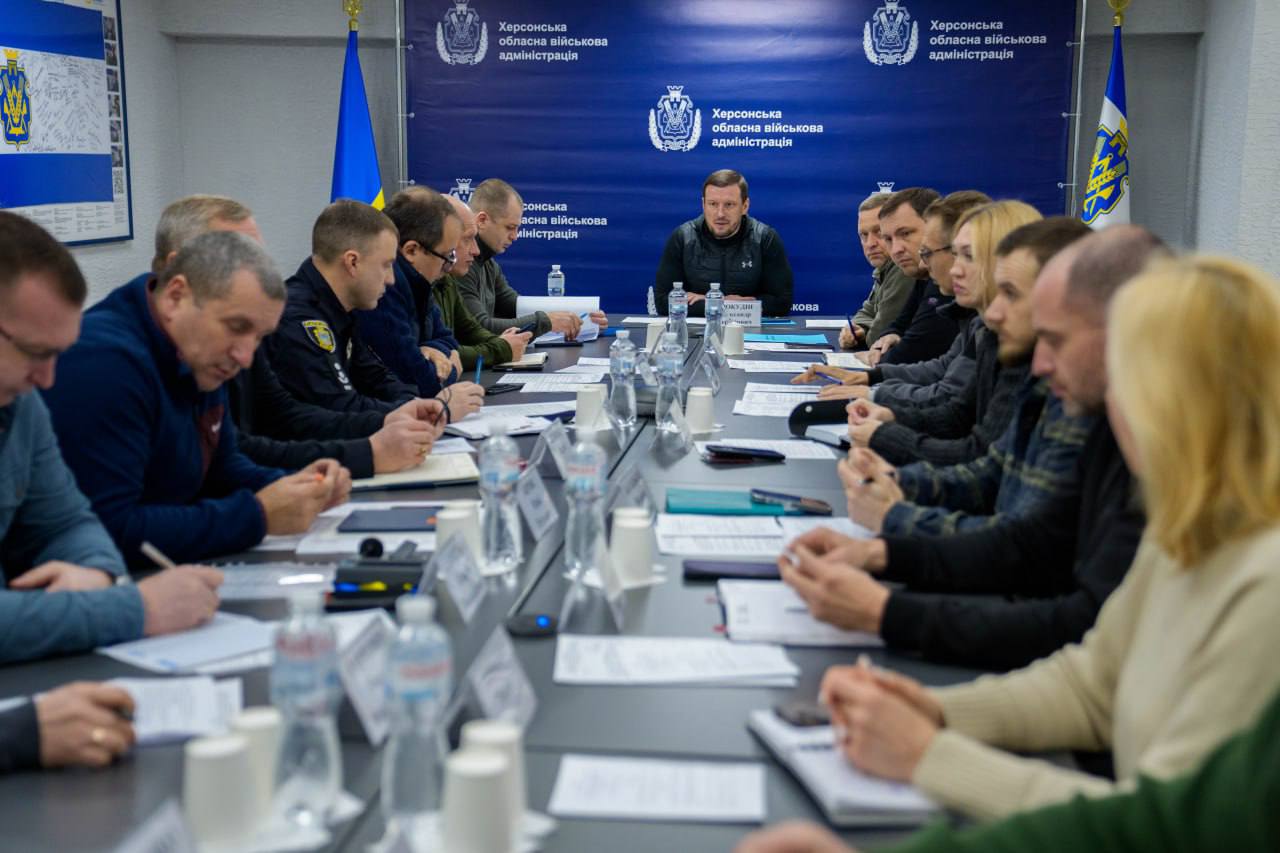 Робоча нарада в Херсонській обласній військовій адміністрації. Група людей у цивільному та офіційному одязі сидить за довгим столом, заставленим паперами та водою. На фоні розташований синій банер із логотипами адміністрації та державний прапор України.