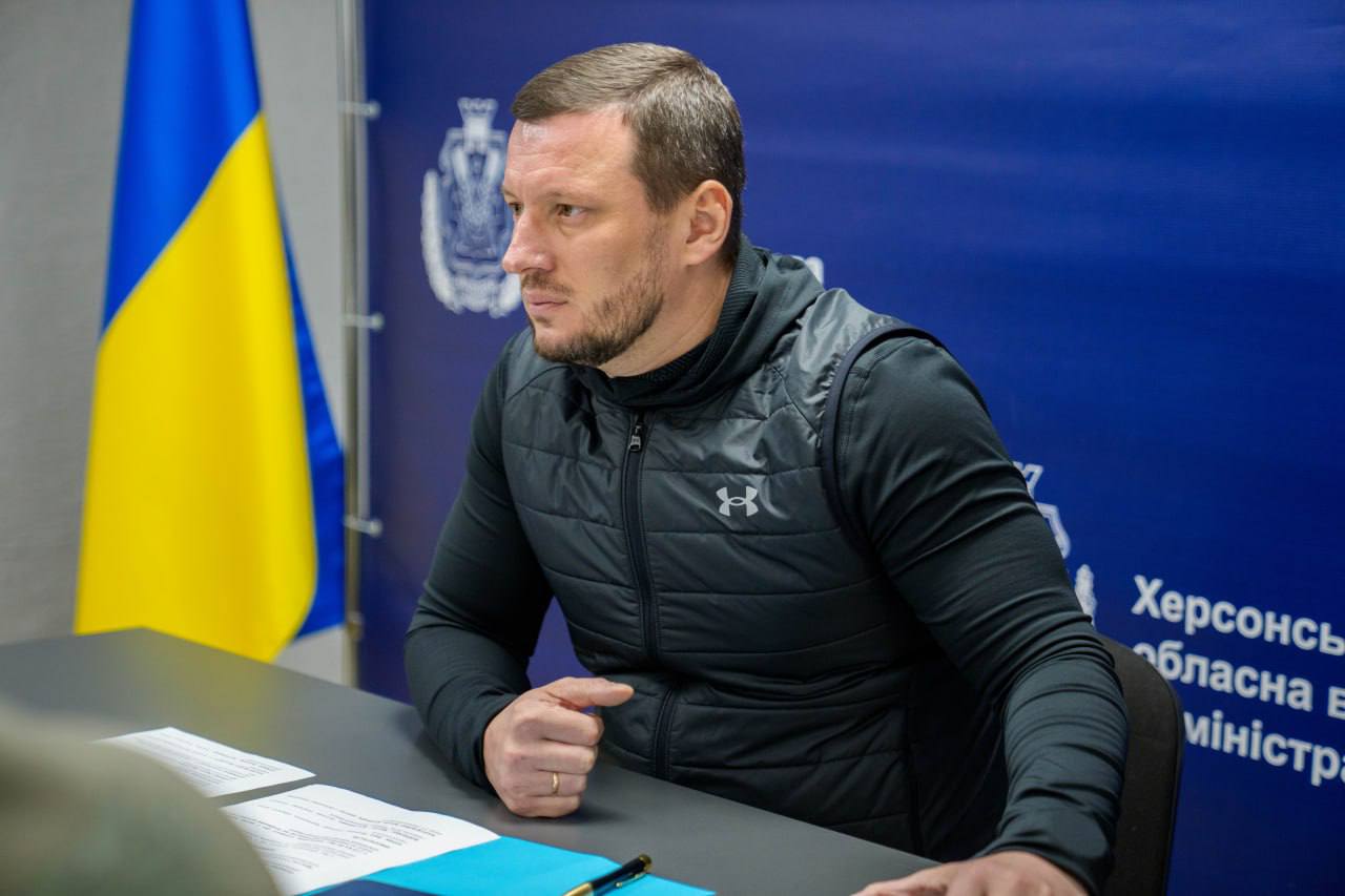 Чоловік у темному спортивному жилеті сидить за столом під час наради. Перед ним лежать робочі документи. На задньому плані — синій банер із логотипом Херсонської обласної військової адміністрації та державний прапор України.