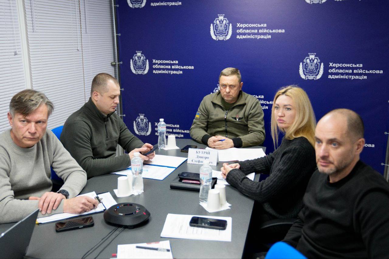 Робоча зустріч групи осіб у конференц-залі. Учасники сидять за столом із документами та пляшками води на фоні синього брендованого банера Херсонської обласної військової адміністрації. Один із присутніх одягнений у куртку кольору хакі з українським прапором на рукаві.