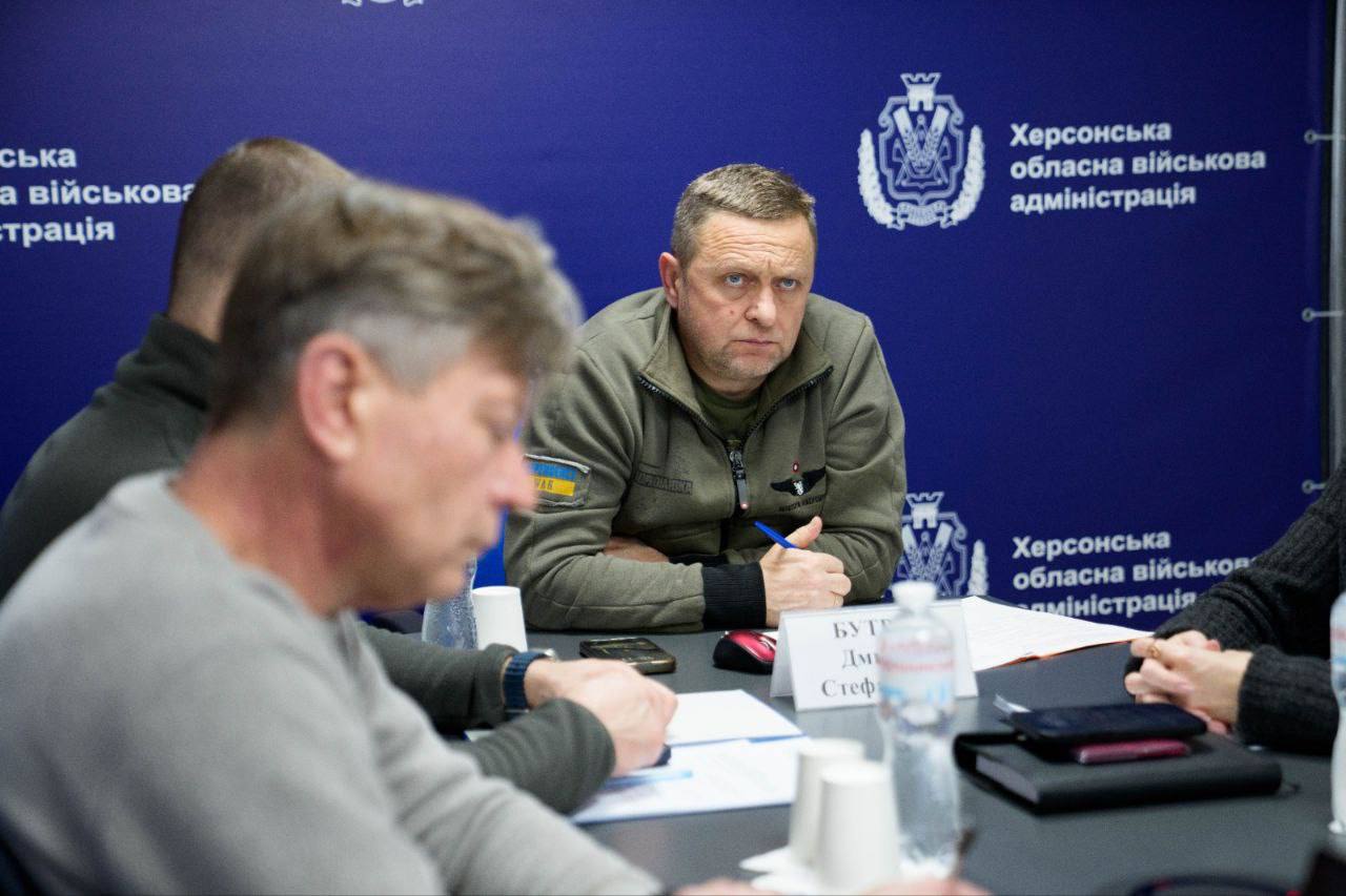 Чоловік у куртці кольору хакі з шевроном у вигляді українського прапора на рукаві сидить за столом під час наради. Він тримає ручку та уважно слухає колег. На задньому плані — синій банер із символікою та написом «Херсонська обласна військова адміністрація».