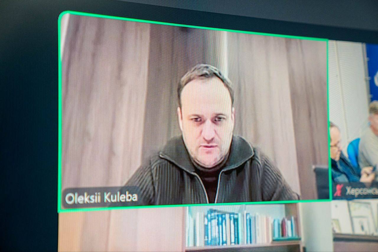 Чоловік у зеленому светрі сидить за робочим столом під час наради. Він тримає ручку та уважно дивиться перед собою. На задньому плані — темно-синій брендований банер із повторюваним логотипом та написом «Херсонська обласна військова адміністрація».