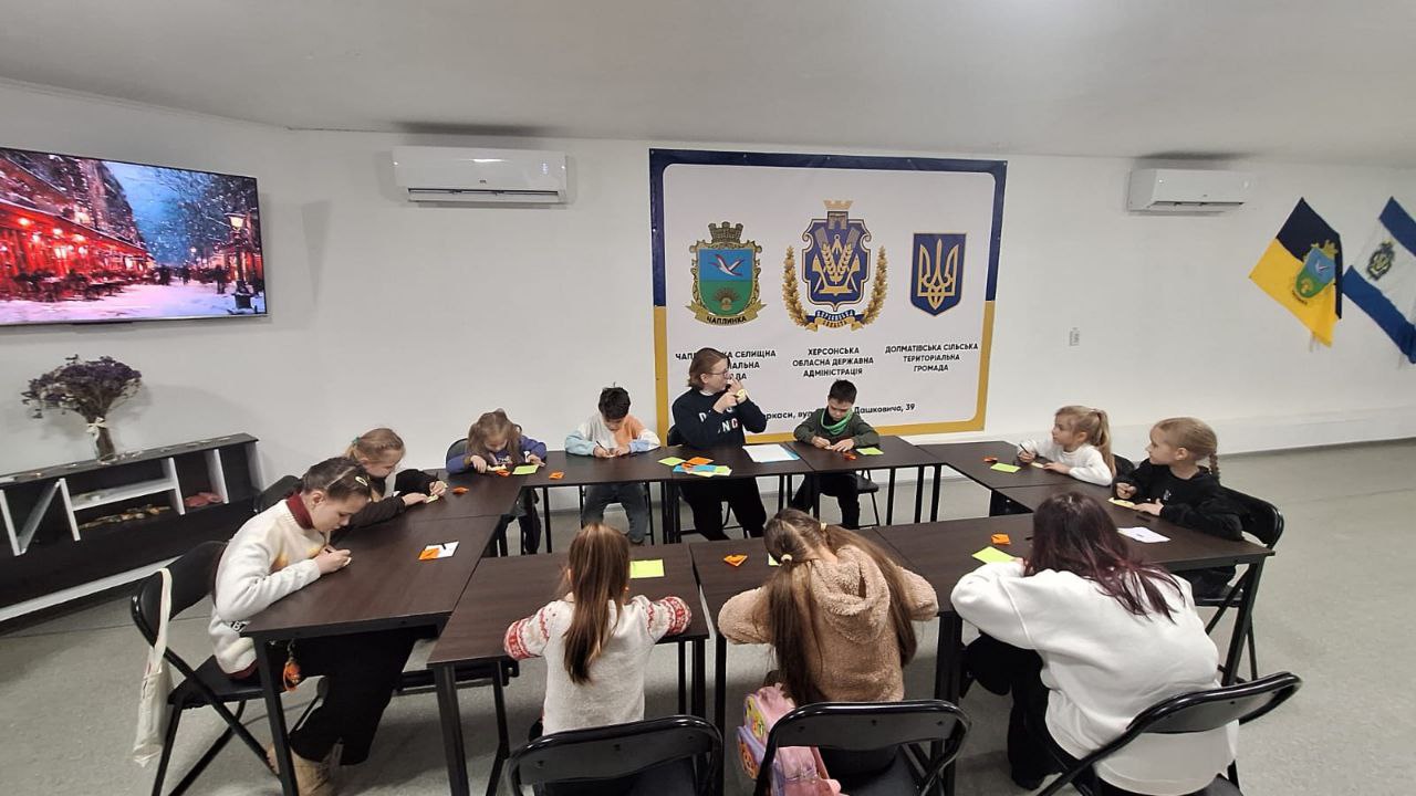 Група дітей та доросла особа сидять за столами, розставленими у формі літери «П», і займаються творчістю. На стіні за ними розташований великий інформаційний стенд із символікою Херсонської області, Чаплинської та Долматівської громад. У приміщенні також видно телевізор, кондиціонери та прапори на стіні.