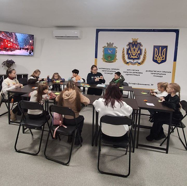 Діти та доросла особа сидять за великим П-подібним столом у навчальному залі та займаються творчістю з папером. На стіні розміщено великий банер із гербами Херсонської області, Чаплинської та Долматівської громад. У куті кімнати працює телевізор, а над банером встановлено кондиціонер.