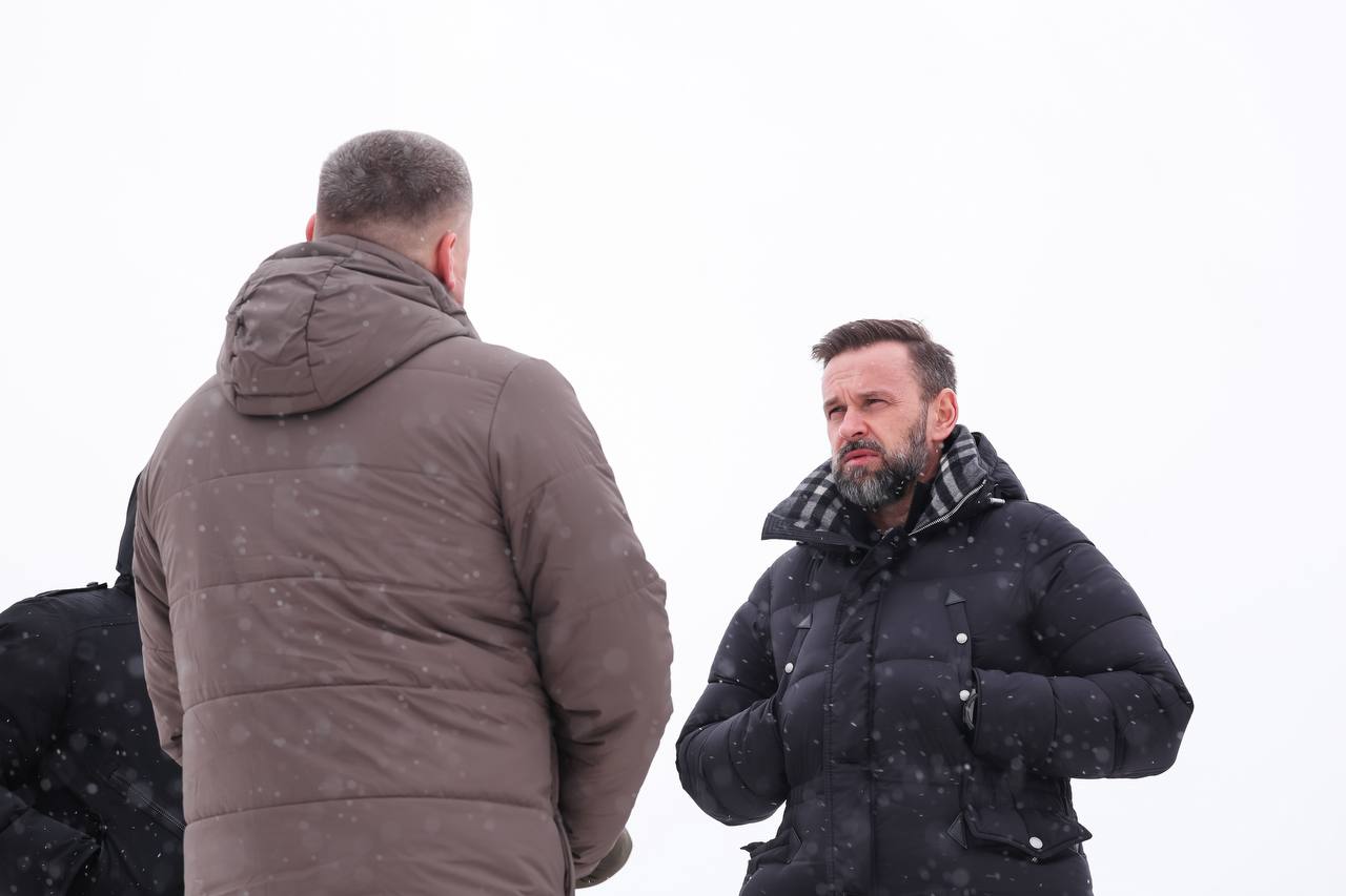 Двоє чоловіків у зимових куртках розмовляють просто неба під час снігопаду. Один стоїть спиною до камери, інший — обличчям.