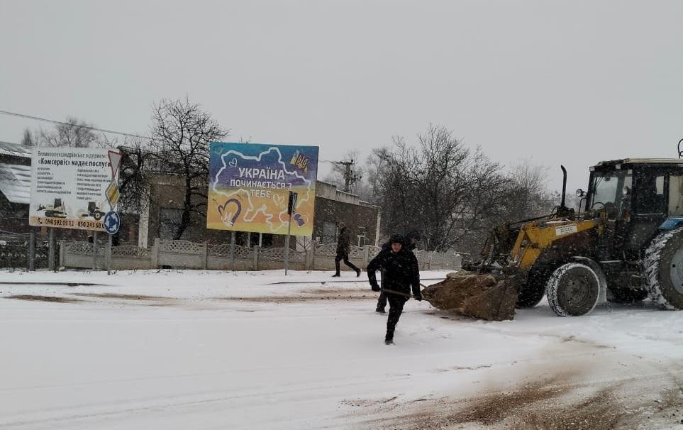 Снігоприбиральна техніка та працівники розчищають засніжену дорогу в населеному пункті. На фоні видно будинки, дерева та патріотичний білборд із написом «Україна починається з тебе».