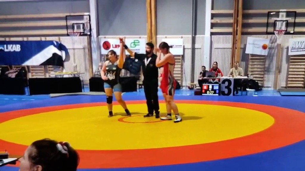 Суддя на борцівському килимі оголошує переможницю змагань, піднімаючи її руку вгору. Поруч стоїть інша спортсменка, на фоні видно трибуни та спортивний зал.