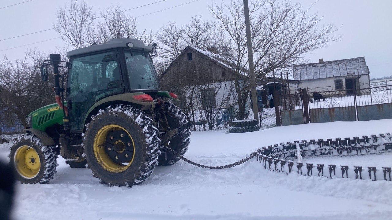 Зелений трактор на ланцюгу тягне металеву конструкцію для розчищення засніженої дороги в селі на фоні приватних будинків.