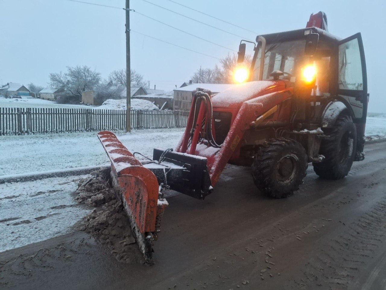 Червоний трактор із піднятими фарами та встановленим відвалом очищає проїжджу частину від мокрого снігу та бруду в сільській місцевості.