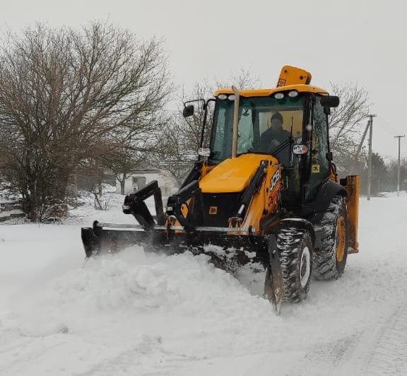 Жовтий екскаватор-навантажувач розчищає дорогу від глибокого снігу в населеному пункті під час снігопаду.